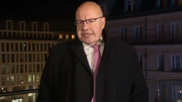 Peter Altmaier looks directly at the camera. He is wearing a white shirt with a pink tie, a black blazer and black overcoat. He is bald and has black-rimmed glasses on.