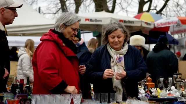 Two woman look at a glass from a stall in a market. One woman in a navy coat holds the glass while the other woman, who is wearing a red coat, looks at the glass.