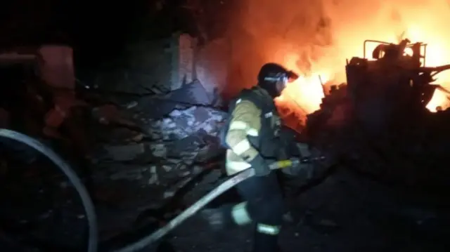 The picture is taken at night. A firefighter walks over rubble to a blaze that glows orange in the dark. There's a lot of smoke. He carries a fire hose and wears a helmet and a fireman uniform