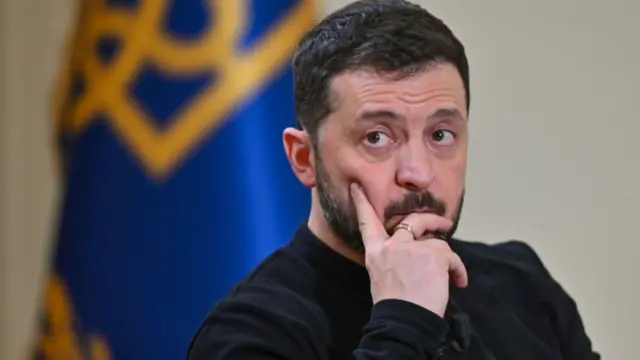 Headshot of Zelensky who looks fed up. He had a hand resting on his chin. He wears a black jumper