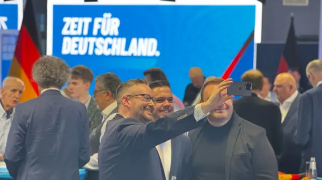 Men smile in suits and take a selfie at the AfD election HQ. A big blue sign is the background.