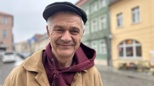 Andreas in a red hoodie and brown coat with houses and a road in the background.