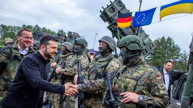 Zelesnky shaking a soldier's hand, as the glad of Ukraine, Germany and the EU flutter in the background.