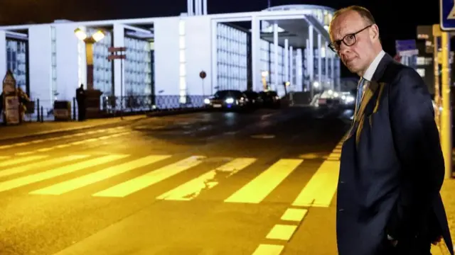 Friedrich Merz looking at the camera as he stands on the edge of a road, waiting to cross it.