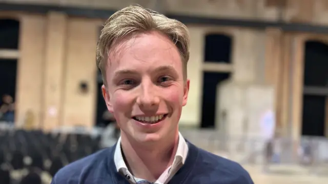 Mika, in a blue jumper with shirt collar, smiling at camera.