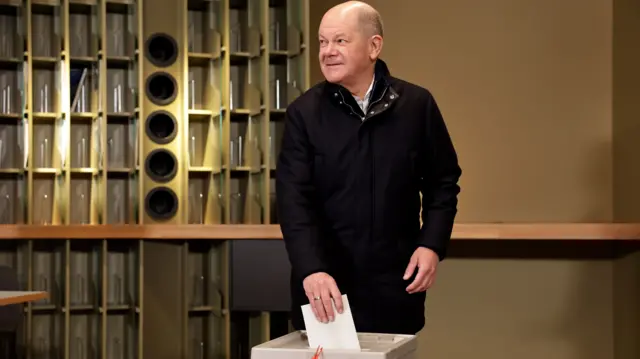 Olaf Scholz poses for photographers as he puts his vote into a white ballot box. He is wearing a black coat.