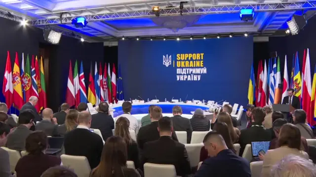 A view of the summit room. People sit in rows of white chairs facing a round table, flanked by a series of flags. The words 'Support Ukraine' appear on a screen at the back of the room.