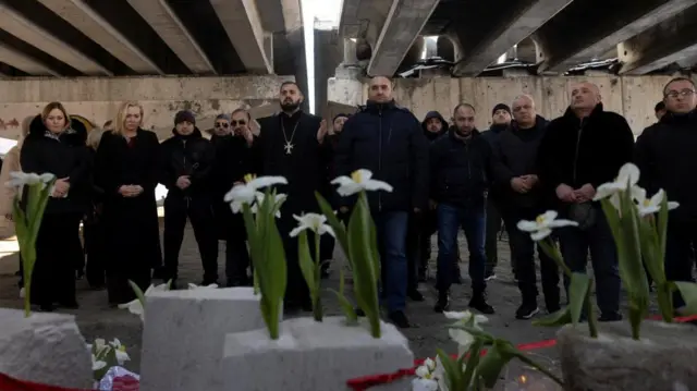 People attend a memorial event marking the third anniversary of Russia's invasion of Ukraine at Romanivka Bridge. A group stands behind memorial flowers.