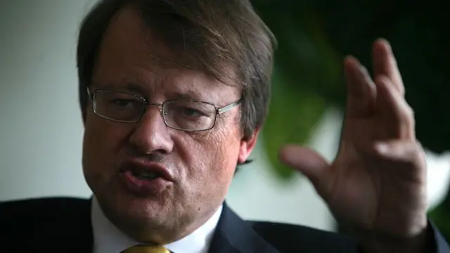 Thomas Matussek speaking in a black suit and yellow tie with his left hand raised