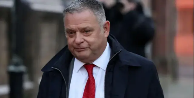 Mike Amesbury MP with gray hair that arrives in court wearing a red tie and a black coat with a zipper