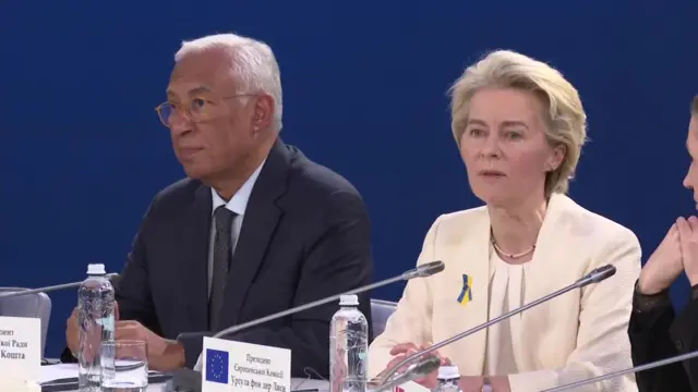 Ursula von der Leyen speaks into a microphone. She wears a cream suit. A yellow and blue ribbon is pinned on her lapel.