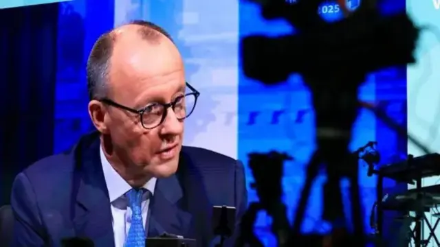 Friedrich Merz speaking to television cameras, wearing a navy suit and a blue tie