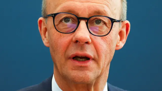 Friedrich Merz wearing a navy suit with small black glasses and a navy background