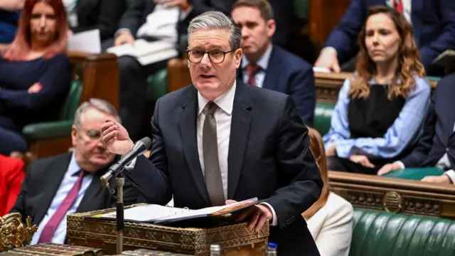 Starmer gestures with his right hand as he stands at the dispatch box, speaking in the House of Commons. Various MPS fill the green benches behind him