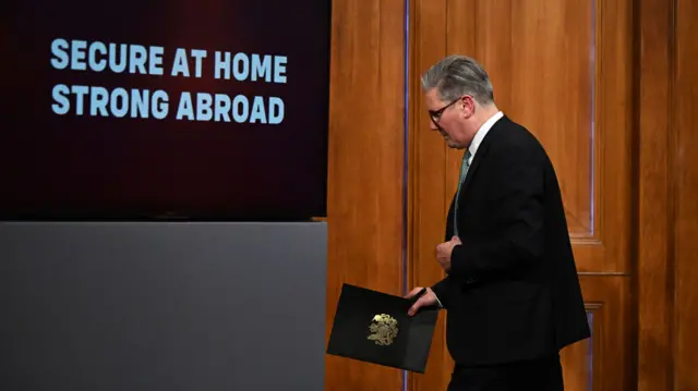 A side view of Starmer as he walks off stage. He holds a folder and a pen and looks down towards the floor. A sign reading 'Secure at Home, Strong Abroad' is in the top left of the image