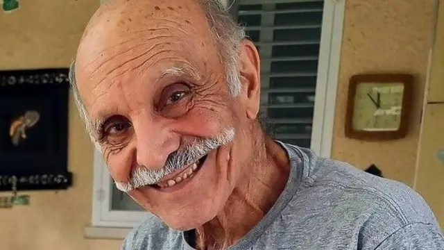 An elderly man with a grey moustache smiles at the camera