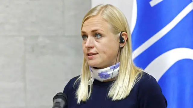 Finland's Foreign Minister Elina Valtonen mid-speech. She is wearing a wired earpiece in her right ear and a white neckerchief with blue patterns. There is a microphone in front of her and in the background, a partial white and blue flag.