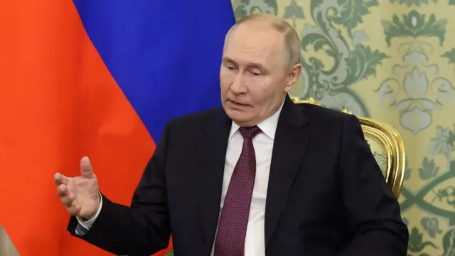 Vladimir Putin sitting on a golden chair with flower-patterned back cushion. He's wearing a black suit, a Russian flag behind him