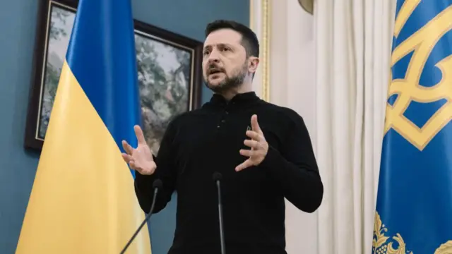 Volodymyr Zelensky delivers speech inside his office, a Ukrainian flag to his right. There's a floral painting in a dark wooden frame hanging on the wall behind him