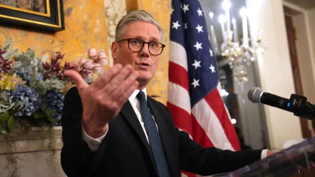 Keir Starmer delivers speech at lectern inside UK ambassador's residence in DC. He's standing in a black suit with a white shirt in front of a marble fireplace with plants and flowers decoration and a US flag to his left