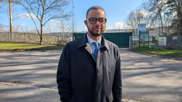 A man with glasses, a long coat, a white shirt and blue tie, stands outside the entrance to a landfill site.