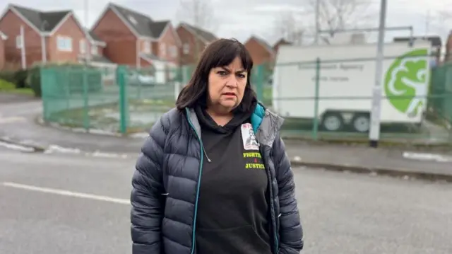 A woman with long black hair and wearing a black hooded jumper and a blue jacket, stands on a road with a green fence and a housing estate behind her.