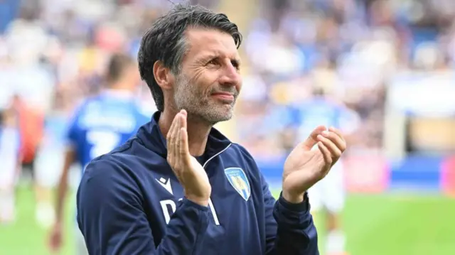 Danny Cowley is smiling as he claps fans. He's wearing Colchester United clothing