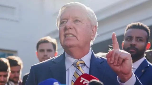 Lindsay Graham beind two microphones with his right hand and finger raised