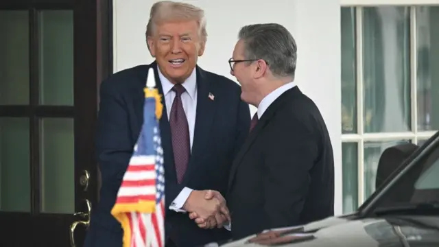 Trump and Starmer shake hands outside the White House. Both men are standing outside the building in suits, smiling. A black car with a US flag on the bonnet partially visible in front of them