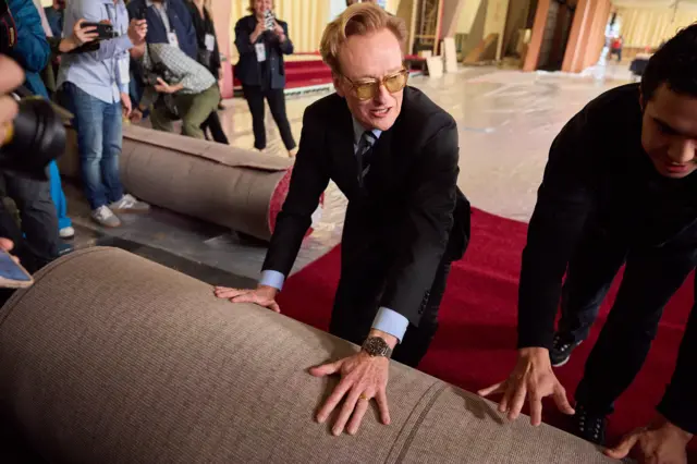 Oscars host Conan O'Brien knelt rolling out the red carpet wearing orange glasses and a black suit