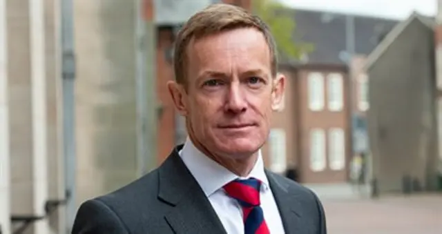 A man with short brown hair, wearing a black jacket, white shirt and red and blue tie, stands in the street.