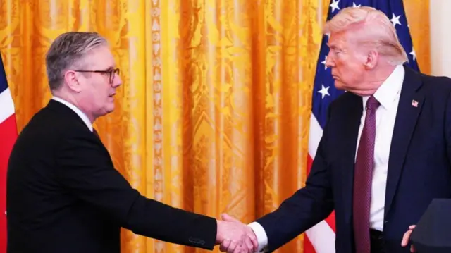 Keir Starmer in a suit shaking hands with Donald Trump, facing each other with orange curtain in background. UK and US flags visible in bakground.