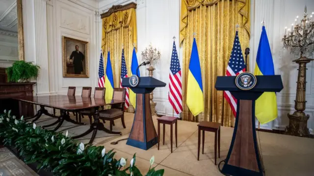 Two podiums and a table at the East Room of the White House, with several US and Ukraine flags in the background.