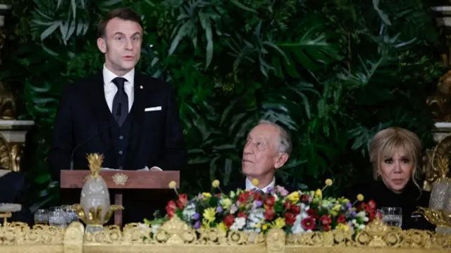 French President Emmanuel Macron standing behind plinth with two people sat at table next to him. Gold detail on table with colourful flowers and green leaves on shrub behind them.