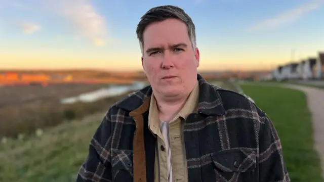 A man standing with a shirt and jacket looking at the camera. He is stood in a field with a path and houses to his left