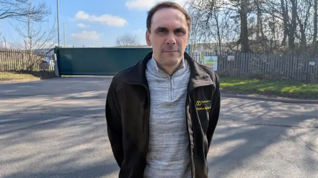 A man wearing a grey top and black jacket stands with his hands behind his back, in front of the entrance to a landfill.