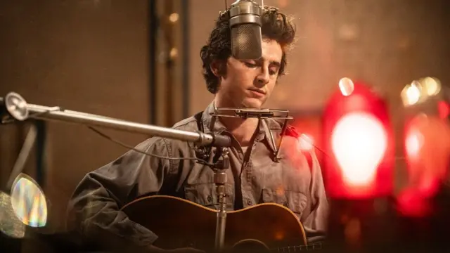 Timothee Chalamet playing Bob Dylan playing the guitar with a harmonica attached