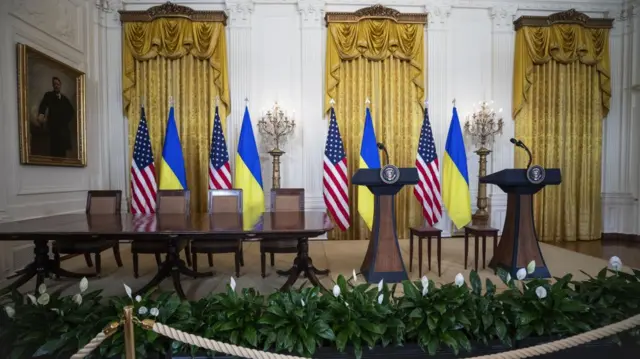 Two podiums and a table at the East Room of the White House, with several US and Ukraine flags in the background.