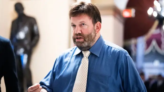 Frank Luntz in a blue shirt and white tie. Looking to the side of the camera with hand slightly raised. Blurred background of statue and grand hall.