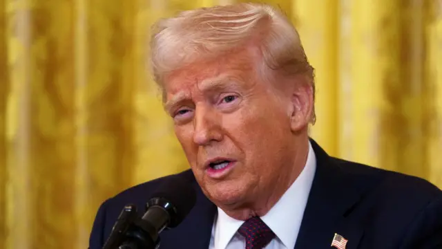 Donald Trump close up as he speaks into a mic, the photo cuts off just below the shoulders. He's wearing a dark blue suit and white shirt with red polka dotted tie. A yellow brocade curtain in the background.