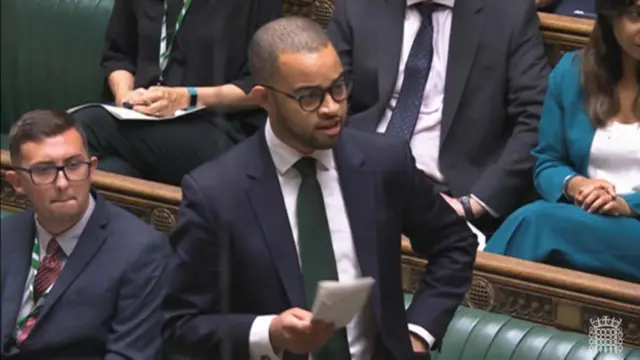 MP Adam Jogee wearing a dark green tie and dark blue suit jacket with a white shirt, stands in the House of Commons to speak while holding a piece of paper in his right hand