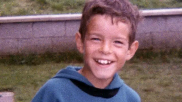 A smiling Shaun McLaughlin stands in front of a wall. He is wearing a blue sweatshirt