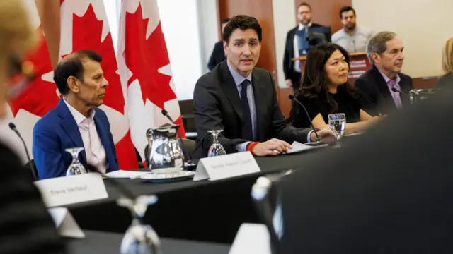 Justin Trudeau sits at a desk with a Canadian flag draped behind him, and several people with serious expressions sitting beside him