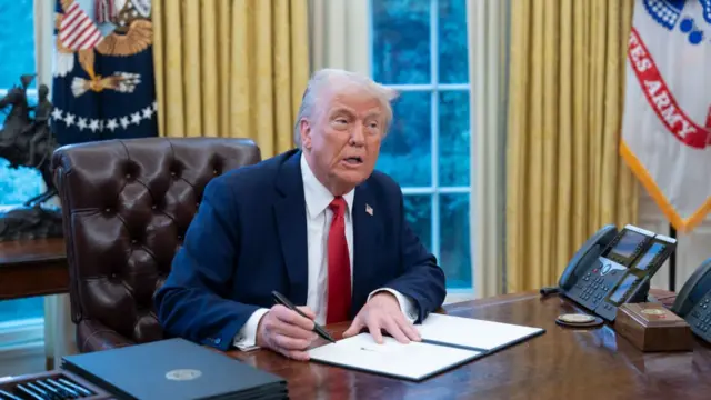 Donald Trump signs a piece of paper at the Resolute Desk in the Oval Office