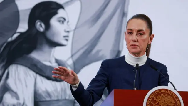 President of Mexico speaks at a podium and gestures with her hand. She is wearing a blue suit jacket.