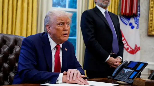 Donald Trump sits inside the Oval Office at a desk and holds a large sharpie pen, as he prepares to sign a document