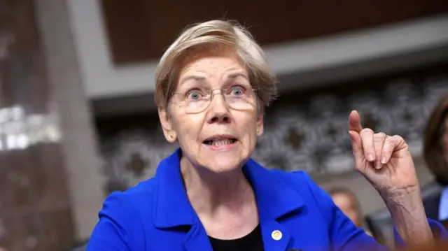 Senator Elizabeth Warren gesticulates with her hand while speaking in Congress. She has short, blonde hair and wears a blue jacket.