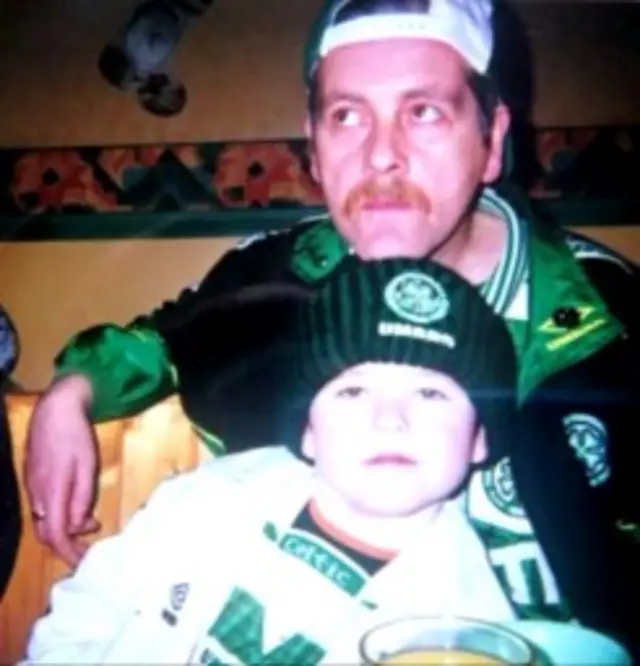 A man wearing a green and black Celtic FC jacket with a green and white Celtic FC top underneath. He is wearing a white cap backwards and has a brown moustache. In front of him is a young boy wearing a white jacket and a green and white Celtic FC top. He is wearing a black woolly hat that has a Celtic FC badge with UMBRO in white letters below it.