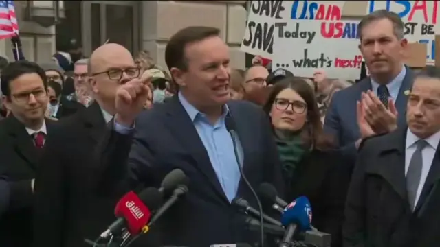 Senator Chris Murphy of Connecticut wears a blue suit without a tie. He stands outside the USAID building with a group of lawmakers and protesters around him.