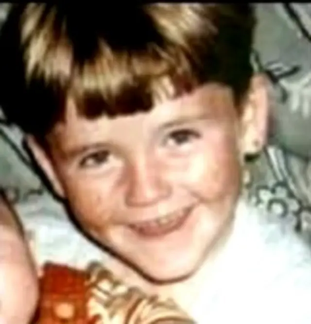 A young boy smiling at the camera, he has brown hair and is wearing a white top